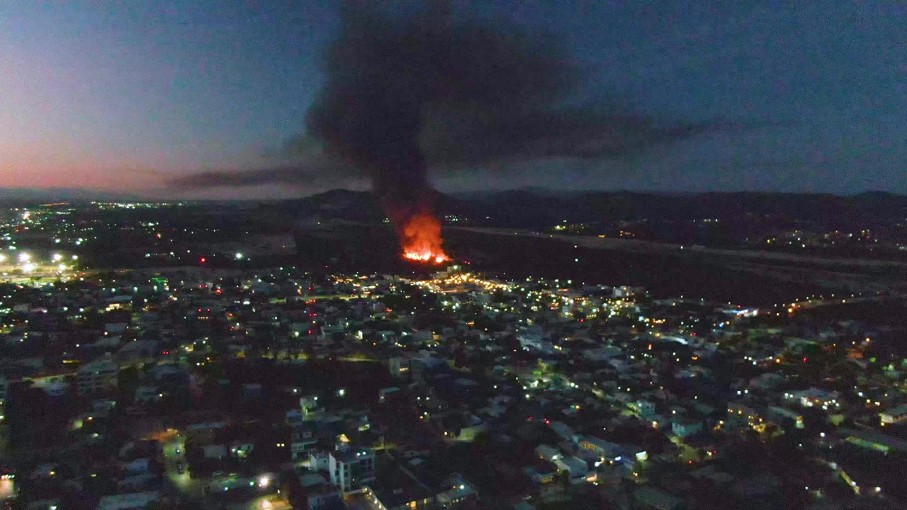 From above San Jose del Cabo, the lower frame is filled with dark buildings and bright street lights while the fire burns near the horizon under a heavy smoke plume, with no visible people.