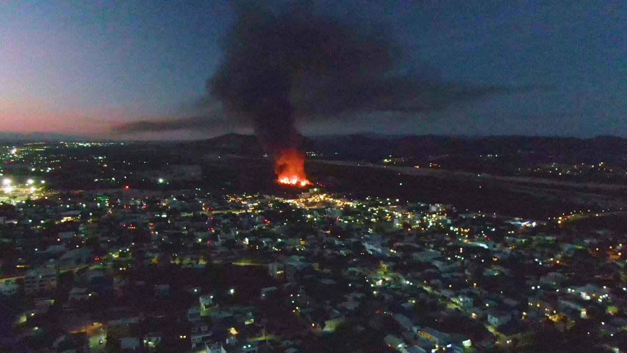 Aerial dusk view over San Jose Del Cabo with city lights below, orange flames in the distance, a black smoke plume rising, and no visible people.