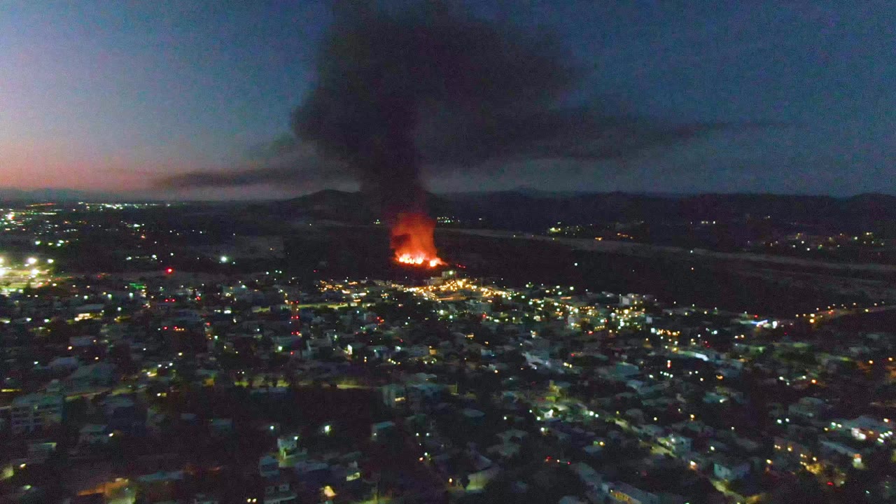 Aerial view over San Jose Del Cabo at dusk with the fire glowing near the center, scattered street lights below, hills on the horizon, and no visible people.