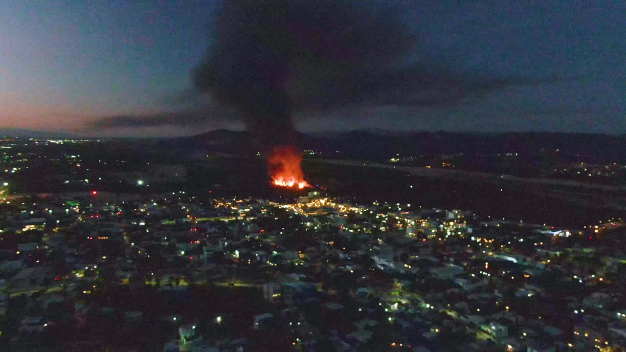 San Jose Del Cabo city lights fill the lower frame as distant flames burn brighter beneath a wide black smoke plume, with no visible people.