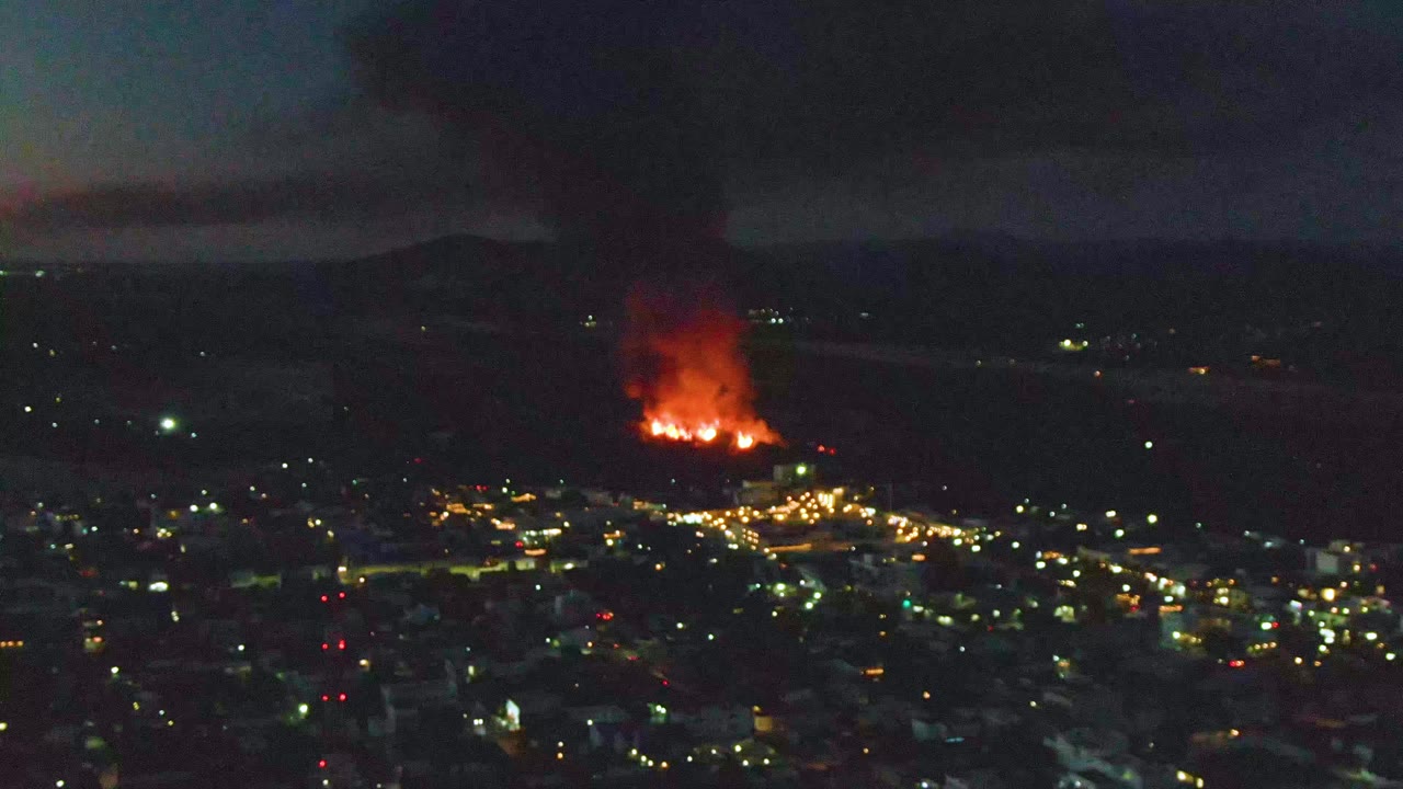San Jose Del Cabo city lights spread across the lower frame while a large fire burns near the horizon and dark smoke rises above it, with no visible people.
