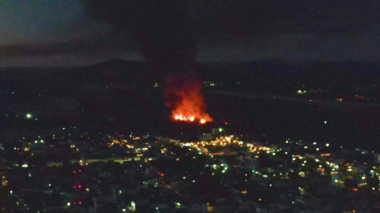 A wide San Jose Del Cabo night view shows orange flames beyond the city lights, with a thick smoke column climbing into the dark sky and no visible people.