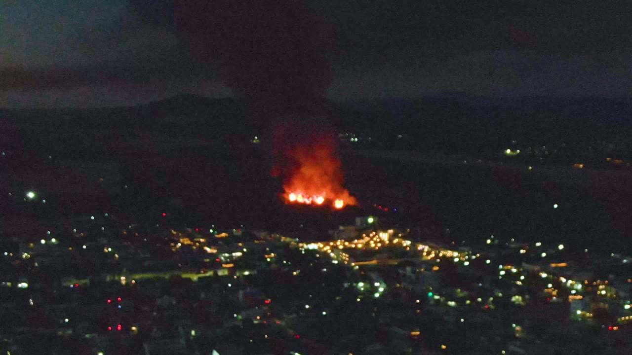 San Jose Del Cabo appears as a dark aerial cityscape with scattered lights, a bright fire at the edge of town, heavy smoke overhead, and no visible people.