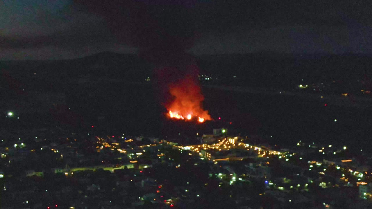 A bright cluster of flames sits beyond the city lights in San Jose del Cabo, sending a dark smoke column upward with no visible people in frame.