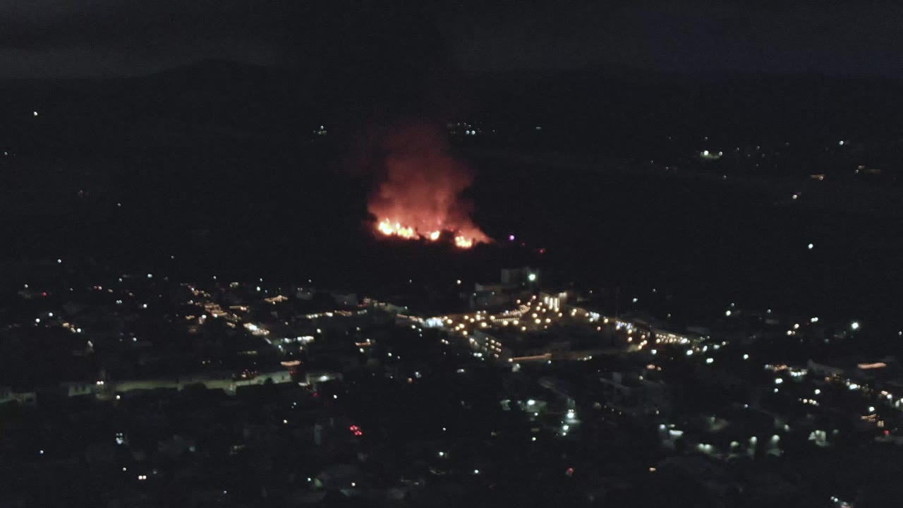 Night aerial city lights with a distant fire and red smoke plume.