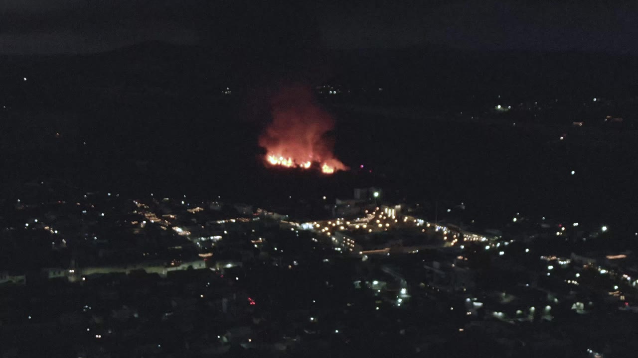San Jose Del Cabo appears as a dark urban grid of lights below a distant orange fire, with red smoke spreading upward and no visible people.