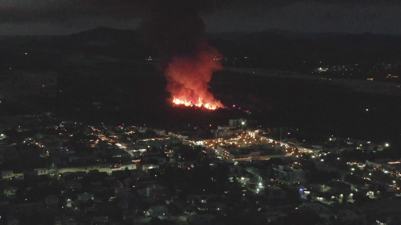 Nighttime city lights with a large fire and smoke plume in the distance.