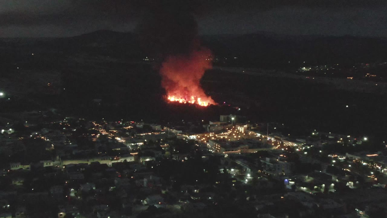 At 0:01, the San Jose del Cabo night aerial holds on a bright fire line at the city edge, with thick smoke above and scattered street lights below, with no visible people.