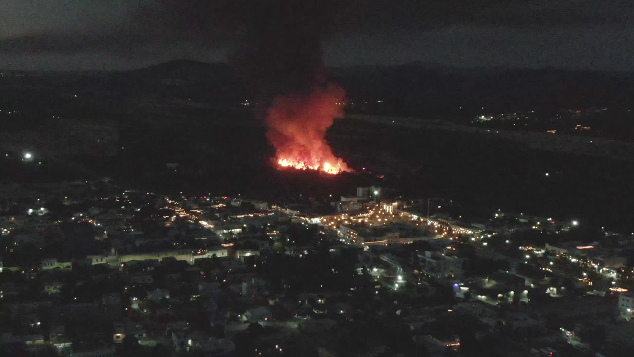 At 0:02, the fire glows red in the midground of San Jose del Cabo while a tall dark smoke plume spreads into the night sky, and no visible people appear in the city below.