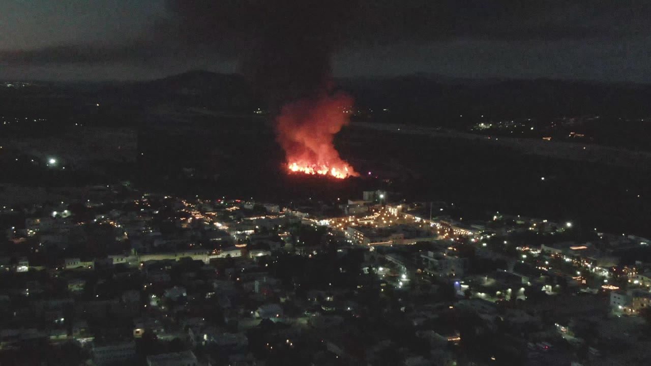 At 0:03, the aerial view over San Jose del Cabo shows dark rooftops in the foreground, bright street lighting in the midground, red flames beyond town, and no visible people.