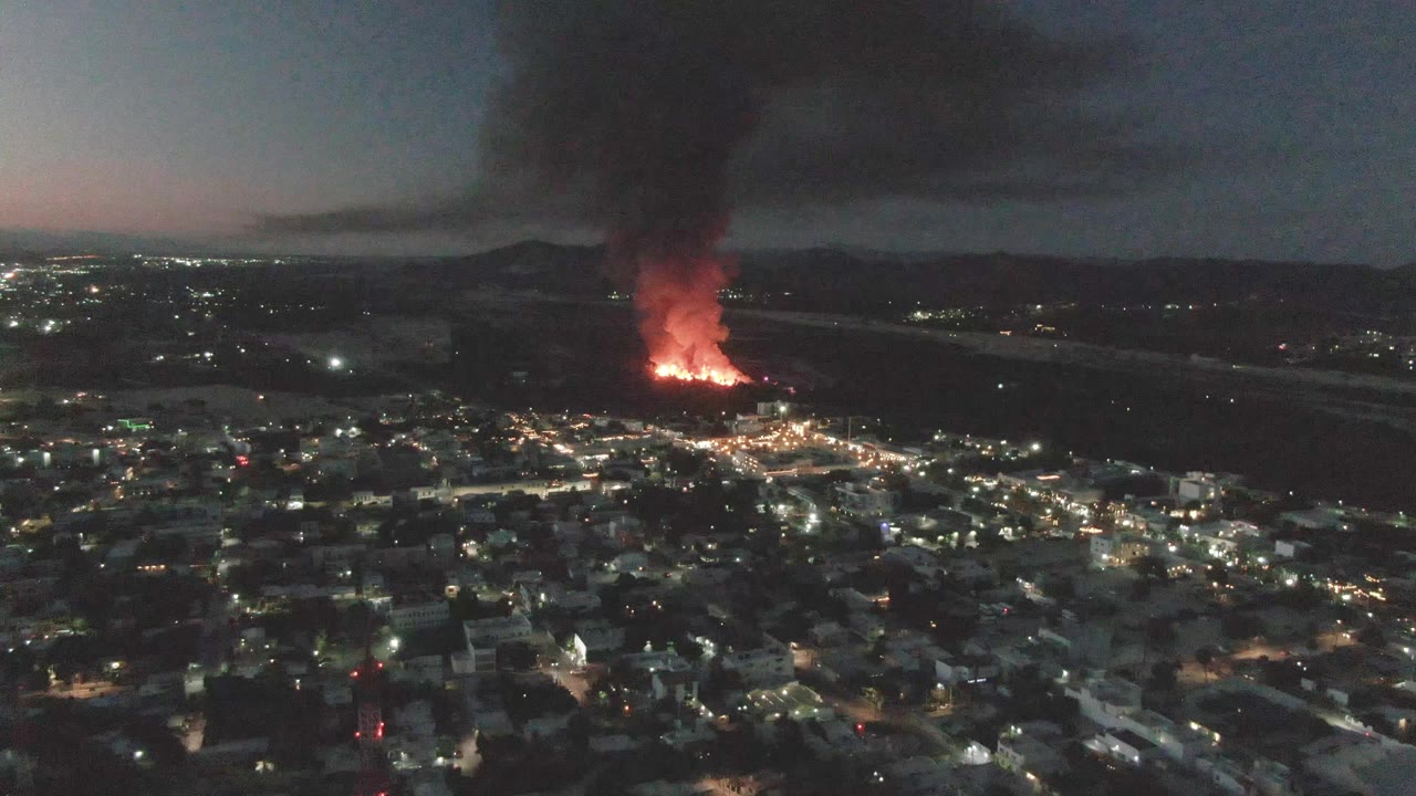 At one second, the San Jose del Cabo city lights remain spread across the lower frame while the red fire glows near the horizon under a dense black smoke plume, with no visible people.