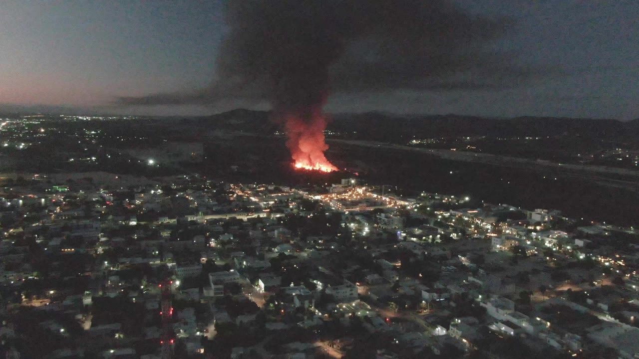 At three seconds, the clip continues from a wide aerial position over San Jose del Cabo, with the fire burning beyond the city lights and black smoke drifting upward into the darkening sky, with no visible people.