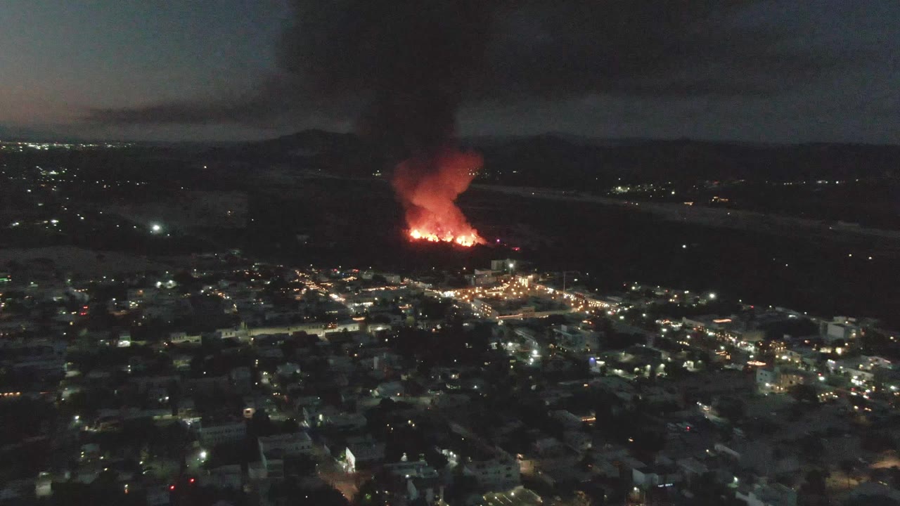 Night aerial view over San Jose del Cabo, Mexico, with red flames and a black smoke column rising beyond lit city blocks and no visible people.
