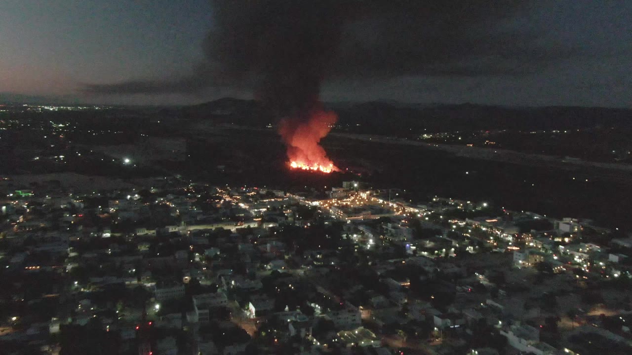Aerial frame of San Jose del Cabo at night with a bright red fire, thick smoke rising upward, scattered city lights, and no visible people.