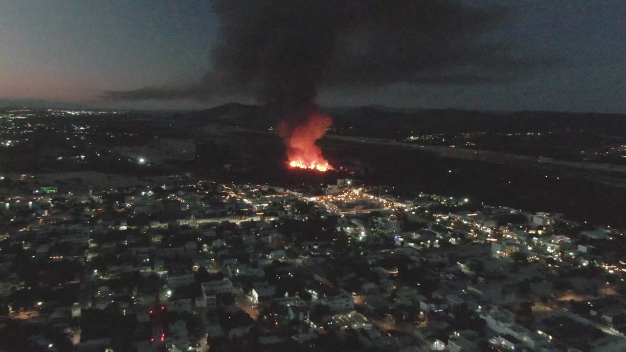 Night drone view over San Jose del Cabo, Mexico, with flames at the edge of the city and a heavy smoke plume spreading above the horizon with no visible people.