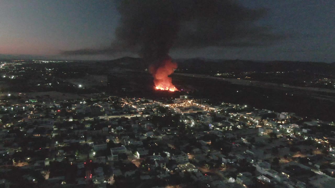 Night city lights below a fire and smoke plume.