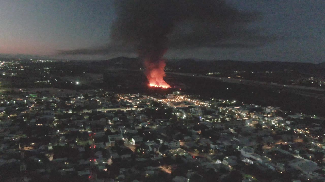 Aerial night view over San Jose del Cabo with city lights in the foreground, a large orange fire in the distance, thick smoke rising, and no visible people.