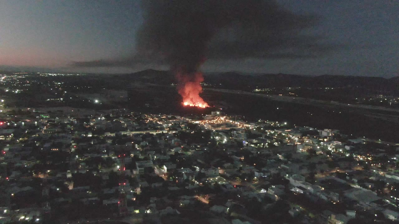 An elevated nighttime view of San Jose del Cabo shows dense city lighting in the lower frame, distant flames near the horizon, a heavy smoke plume above, and no visible people.