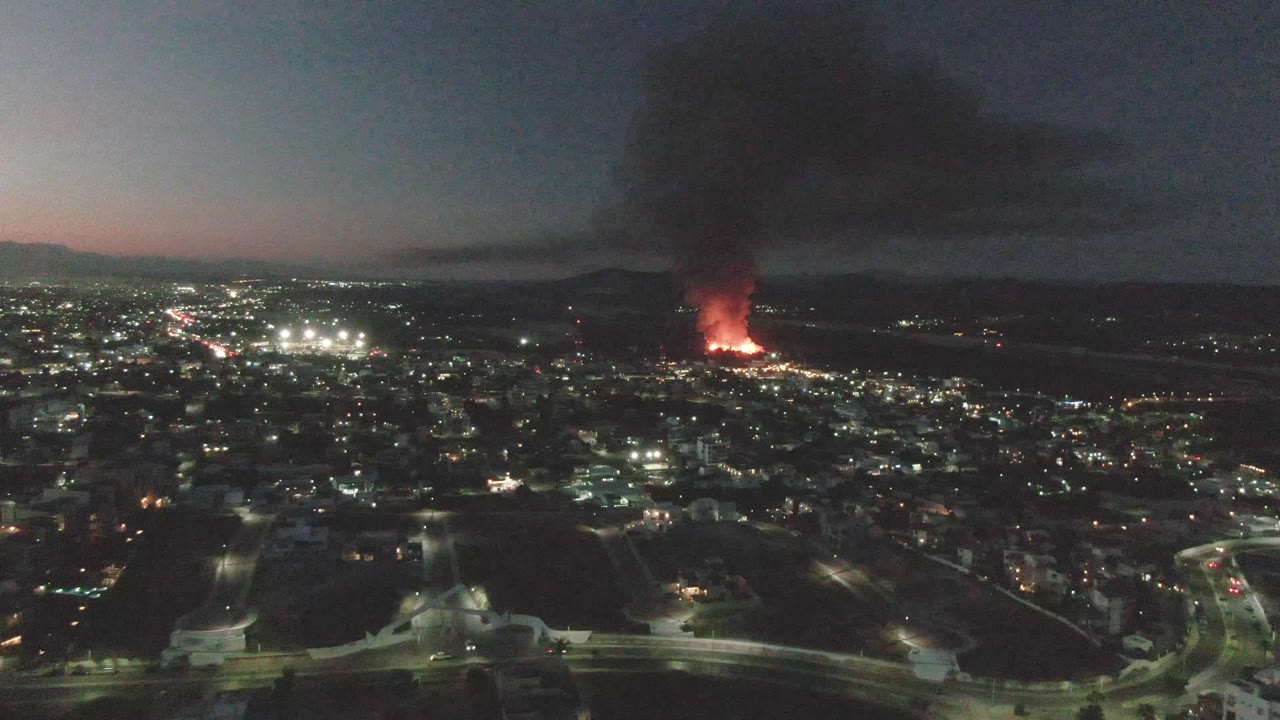 San Jose del Cabo appears from above with illuminated streets in the foreground, clustered city lights, a red fire near the horizon, heavy dark smoke, and no visible people.