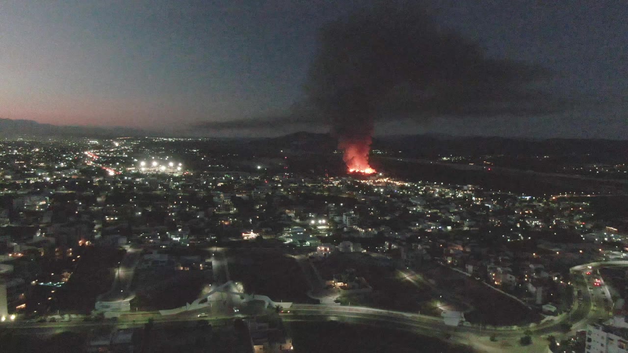 A twilight aerial over San Jose del Cabo shows bright urban lights, curved foreground roads, distant red flames, a thick black smoke plume drifting upward, and no visible people.
