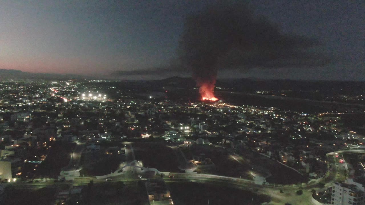 A wide aerial frame over San Jose del Cabo shows foreground streets, scattered buildings, bright city lights, a red-orange fire beyond the urban area, rising black smoke, and no visible people.
