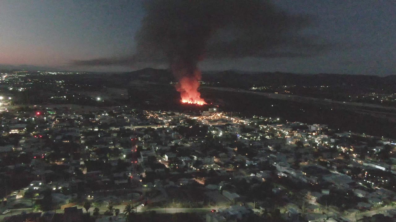 Night aerial view over San Jose Del Cabo, Mexico, with city lights below, distant orange flames near the horizon, a thick black smoke plume, and no visible people.