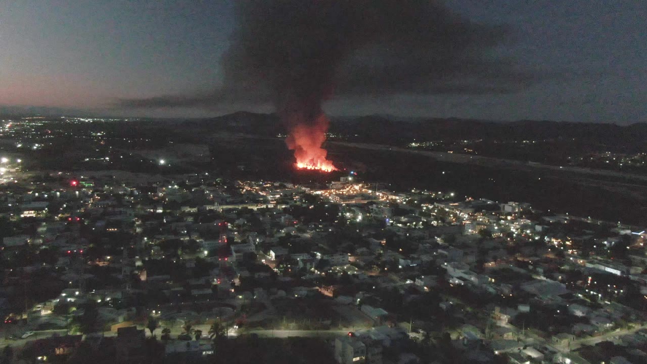 Aerial nighttime frame over San Jose Del Cabo, Mexico, showing a bright fire beyond residential blocks, smoke spreading across the sky, and no visible people.
