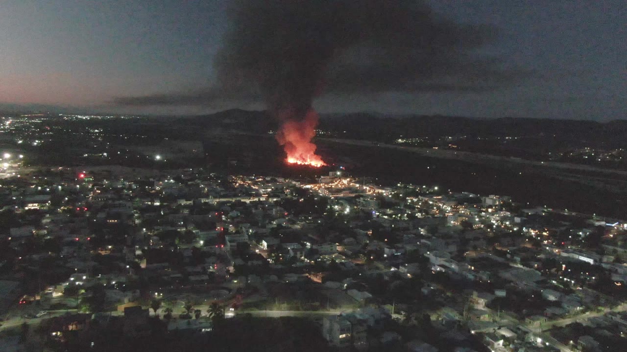 Night drone view in San Jose Del Cabo, Mexico, with scattered city lights in the foreground, orange fire in the distance, black smoke above it, and no visible people.