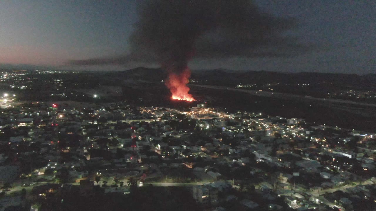 Wide aerial city view with distant flames under a dark sky.