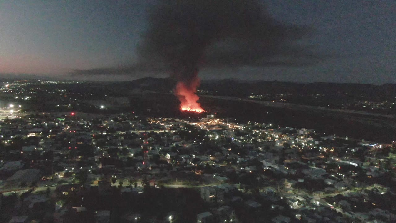 Opening frame over San Jose Del Cabo at night, showing city lights below, a bright orange fire in the distance, a dark smoke plume rising, and no visible people.