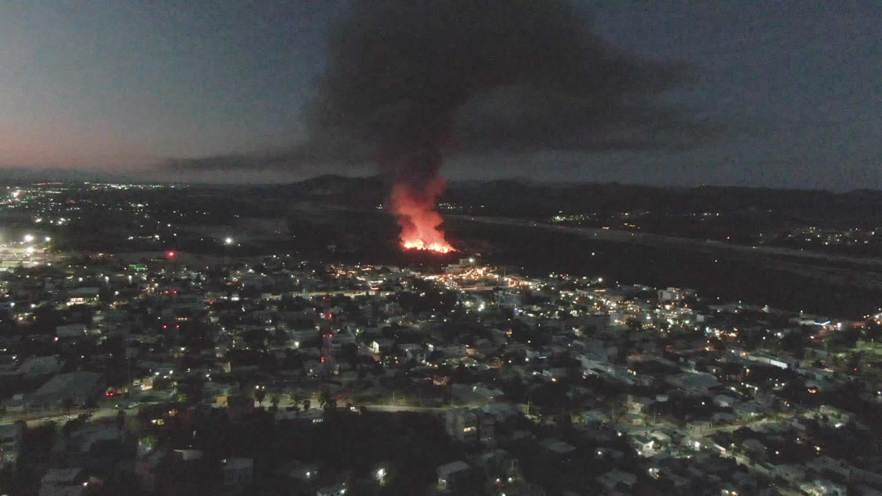 One second into the clip, the San Jose Del Cabo aerial view holds on the glowing fire, dense black smoke above it, scattered city lights, and no visible people.