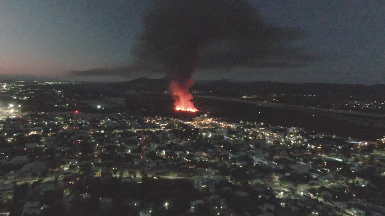 At 0:02, the San Jose Del Cabo city lights remain below a bright fire line, with a tall dark smoke plume broadening over the horizon and no visible people.