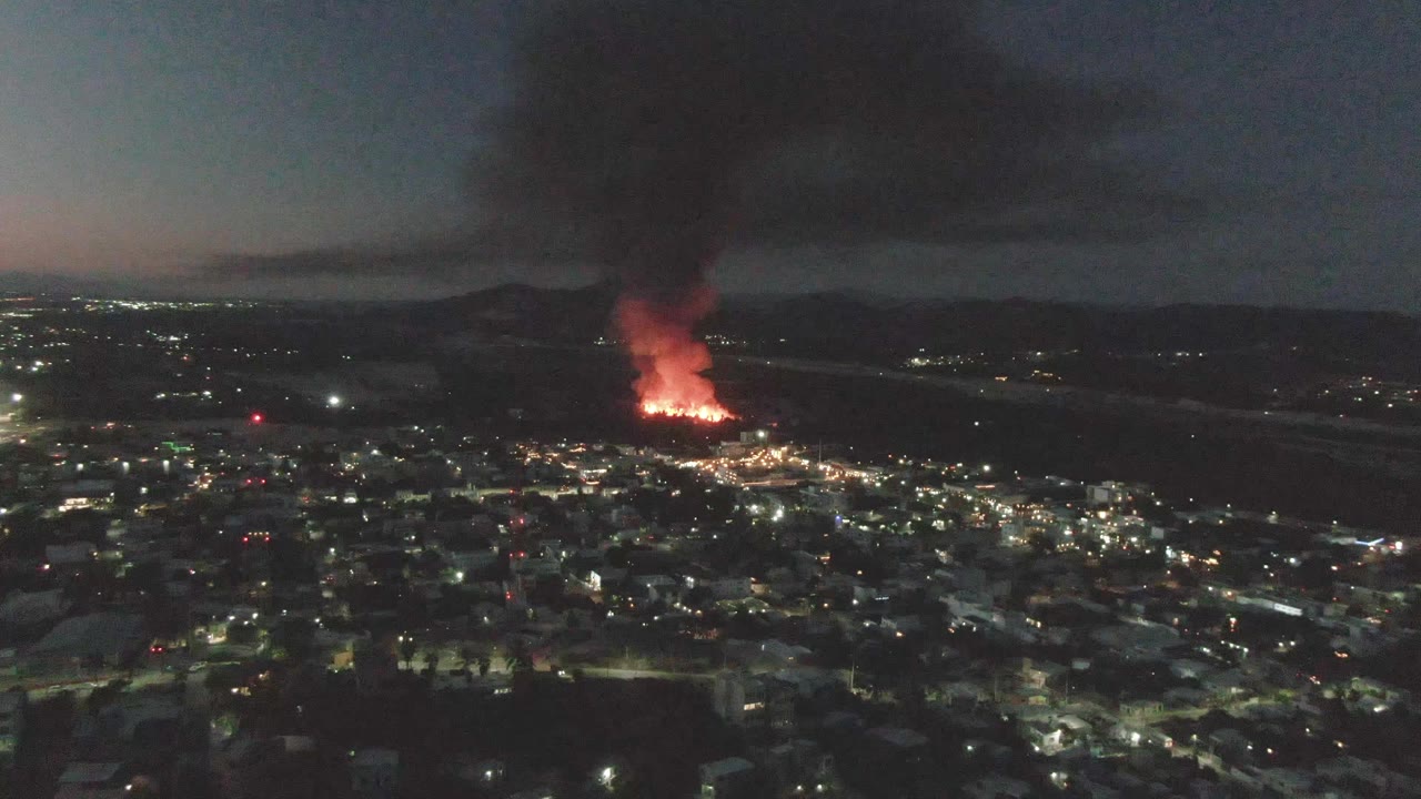 Wide nighttime city aerial with a fire burning beyond the foreground streets.