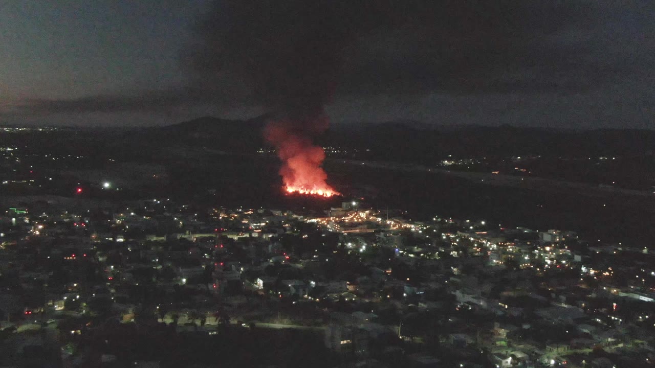 Opening frame of San Jose del Cabo at night, with city lights below, orange flames in the distance, a smoke plume rising into the dark sky, and no visible people.