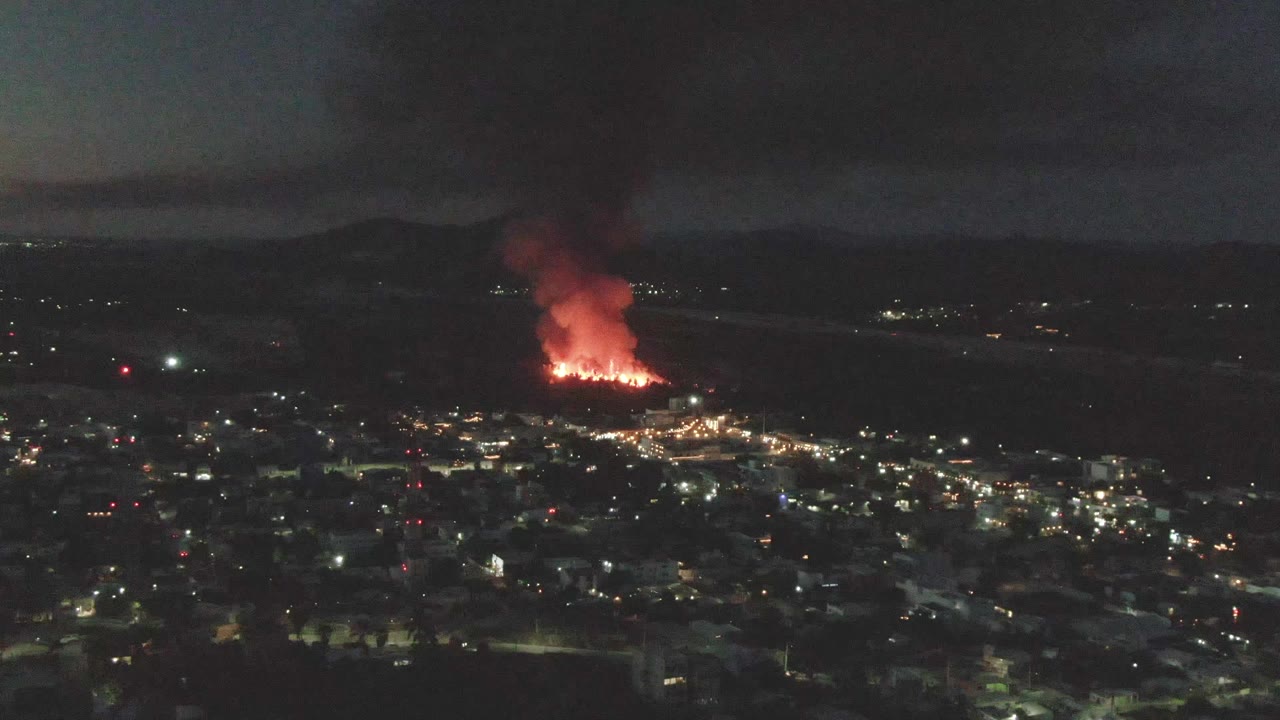 San Jose del Cabo night view one second in, with the fire glowing brighter against the dark hillside area, smoke lifting upward, and no visible people.