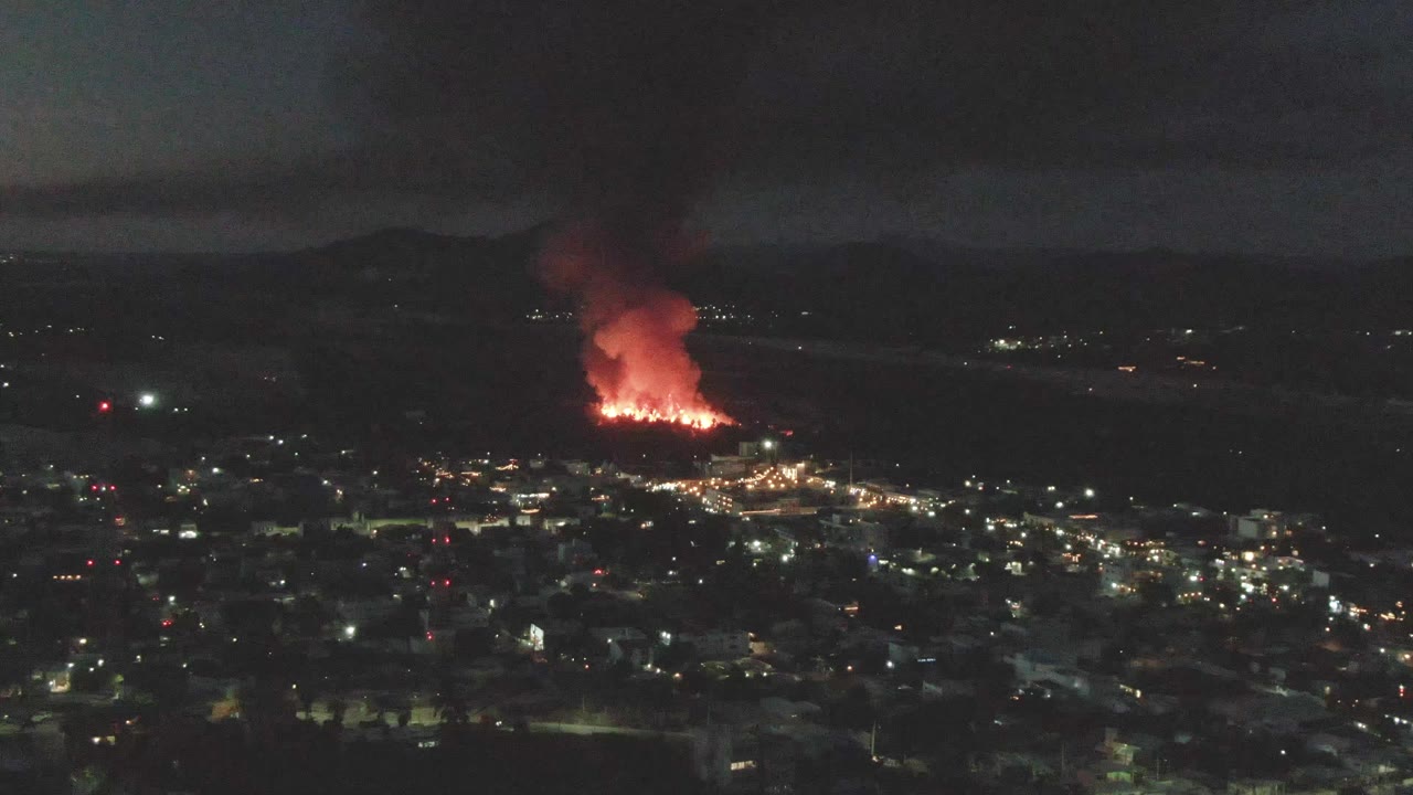 San Jose del Cabo at night with scattered city lights in the foreground, a concentrated orange fire in the distance, smoke spreading upward, and no visible people.