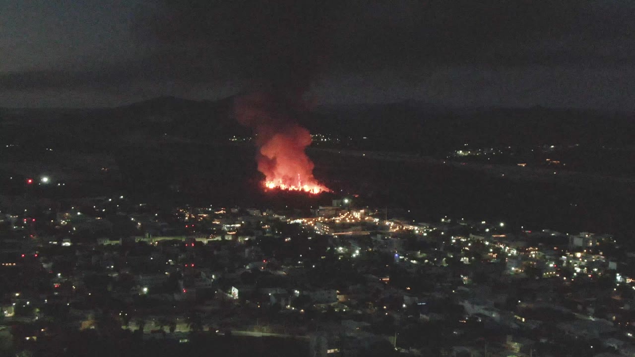 Mid-clip San Jose del Cabo night aerial view with a darker foreground, bright city lights, active flames near the horizon, a thick smoke column, and no visible people.