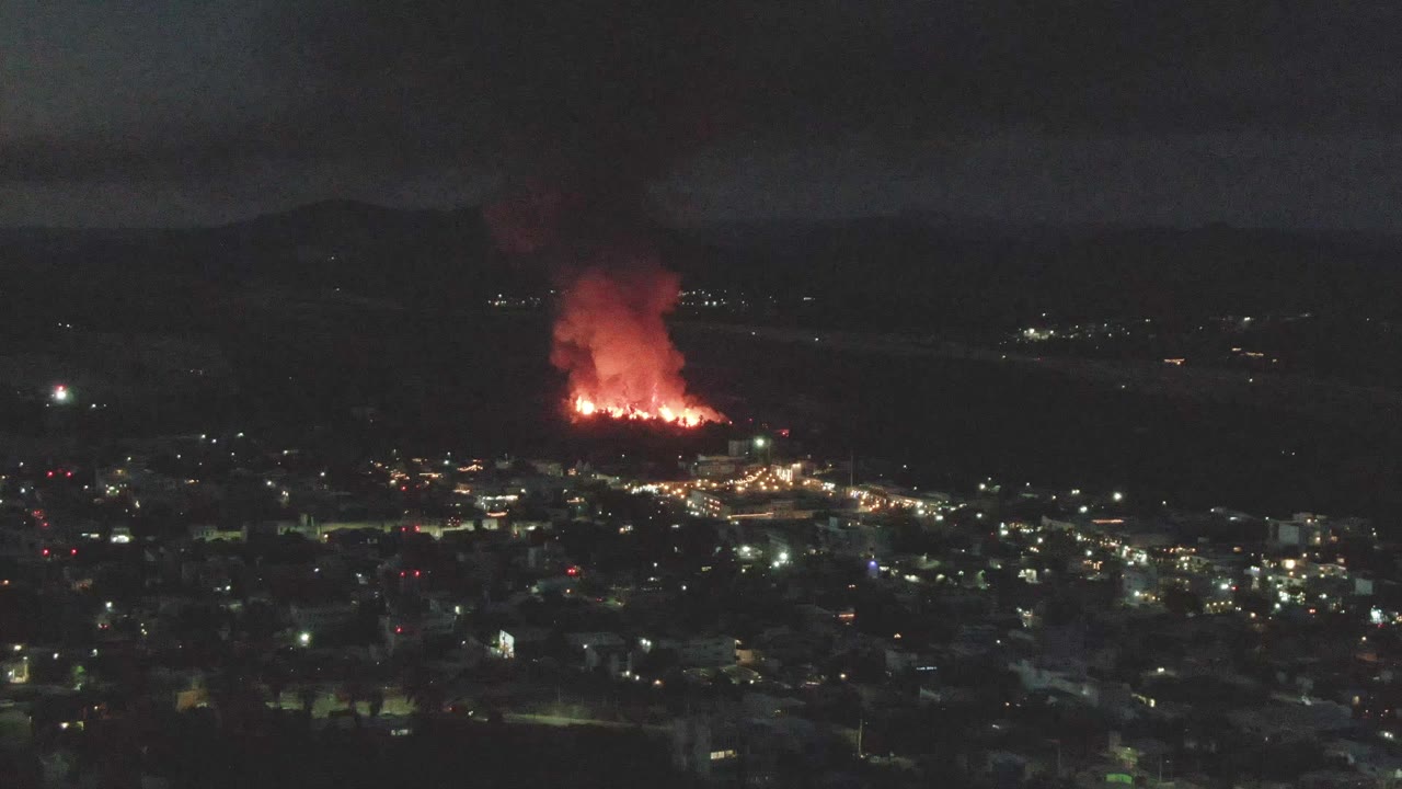 Night cityscape with a distant red fire and smoke plume.