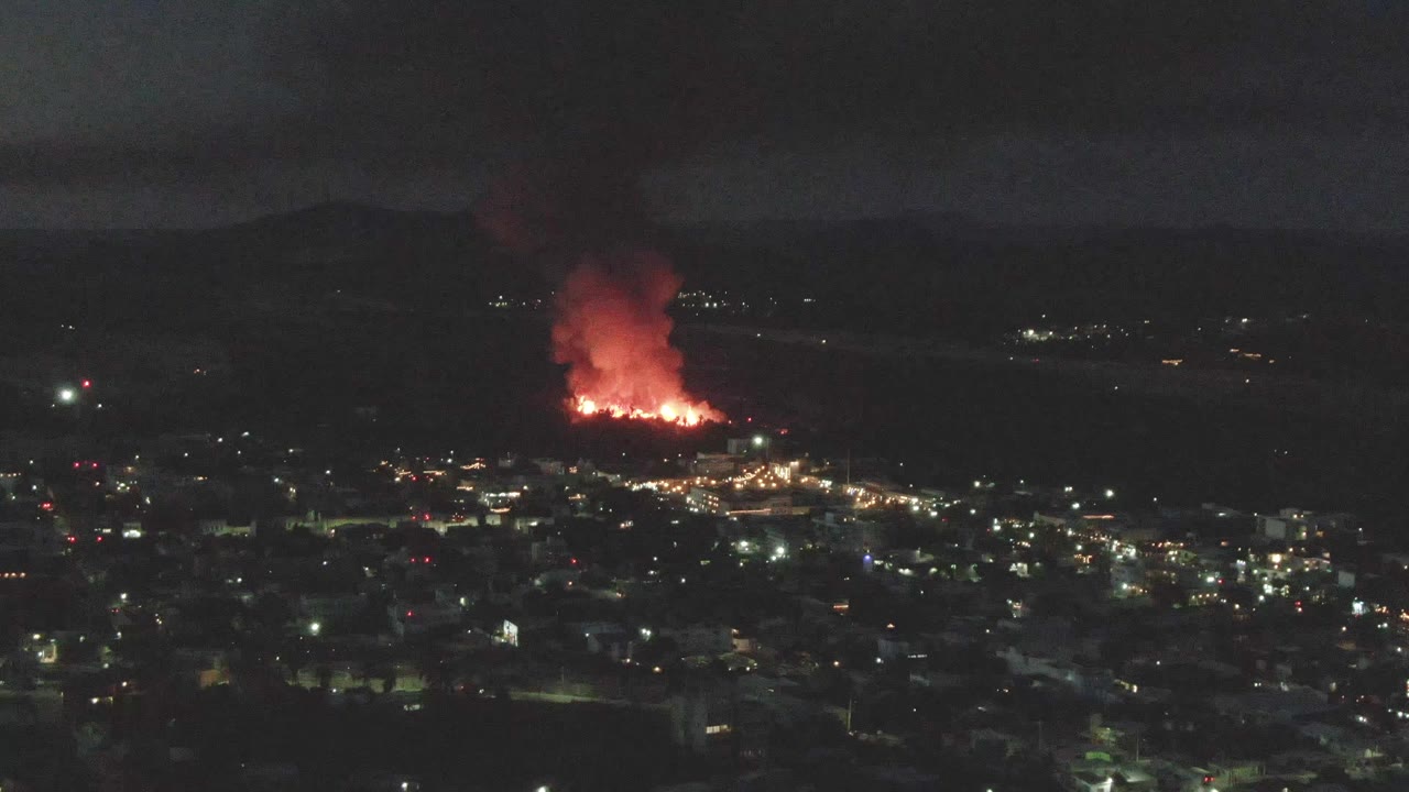 At 0:02, a red-lit smoke plume rises from the fire beyond San Jose del Cabo, with the city lights spread across the lower frame and no visible people.