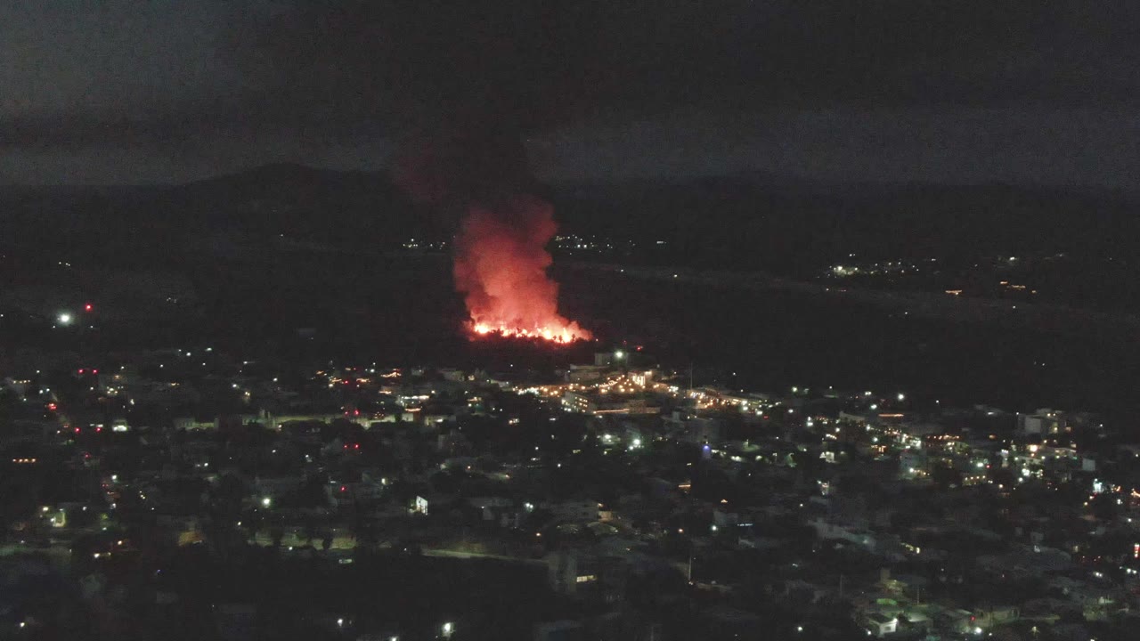At 0:03, the distant fire burns near the left side of the San Jose del Cabo night skyline, with dark terrain in front and no visible people.