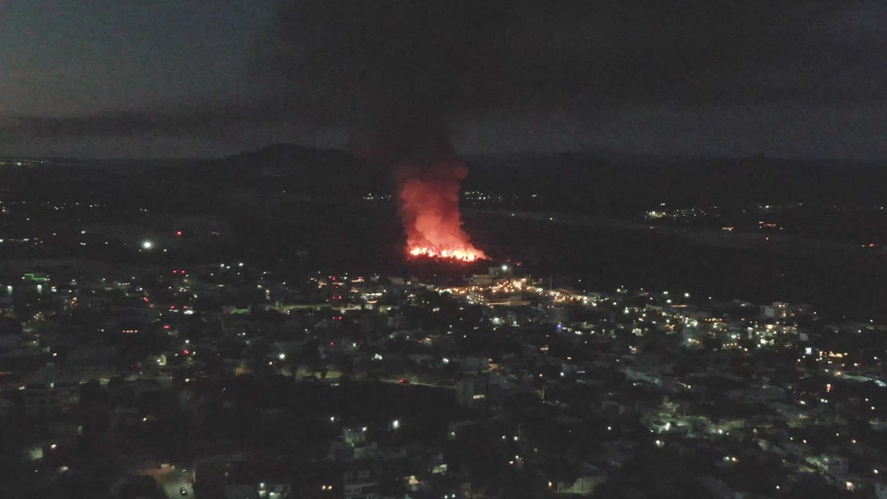 A second nighttime aerial frame over San Jose del Cabo keeps the fire near the horizon, with orange-red flames, dense smoke, scattered city lights, and no visible people.