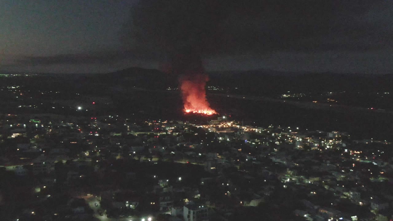 The frame shows San Jose del Cabo at night with a bright fire burning in the distance, a broad black smoke plume above it, dim hills behind it, and no visible people.