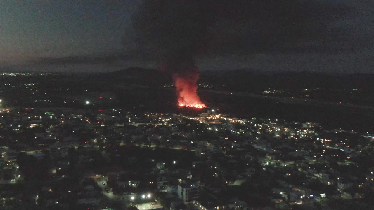 A nighttime aerial view of San Jose del Cabo shows the fire slightly left of center, with dark foreground buildings, scattered lights, a heavy smoke column, and no visible people.