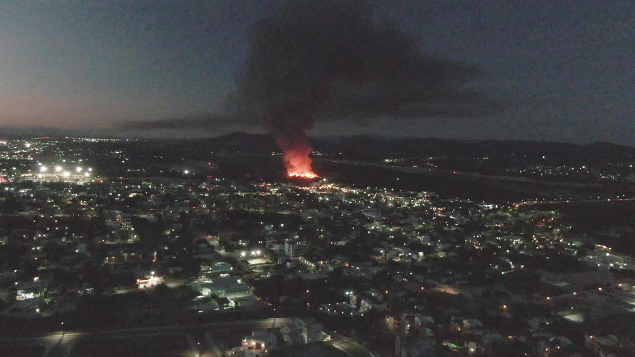 At 0:00, San Jose del Cabo city lights fill the lower frame while a distant red fire sends black smoke into the dark sky, with no visible people.