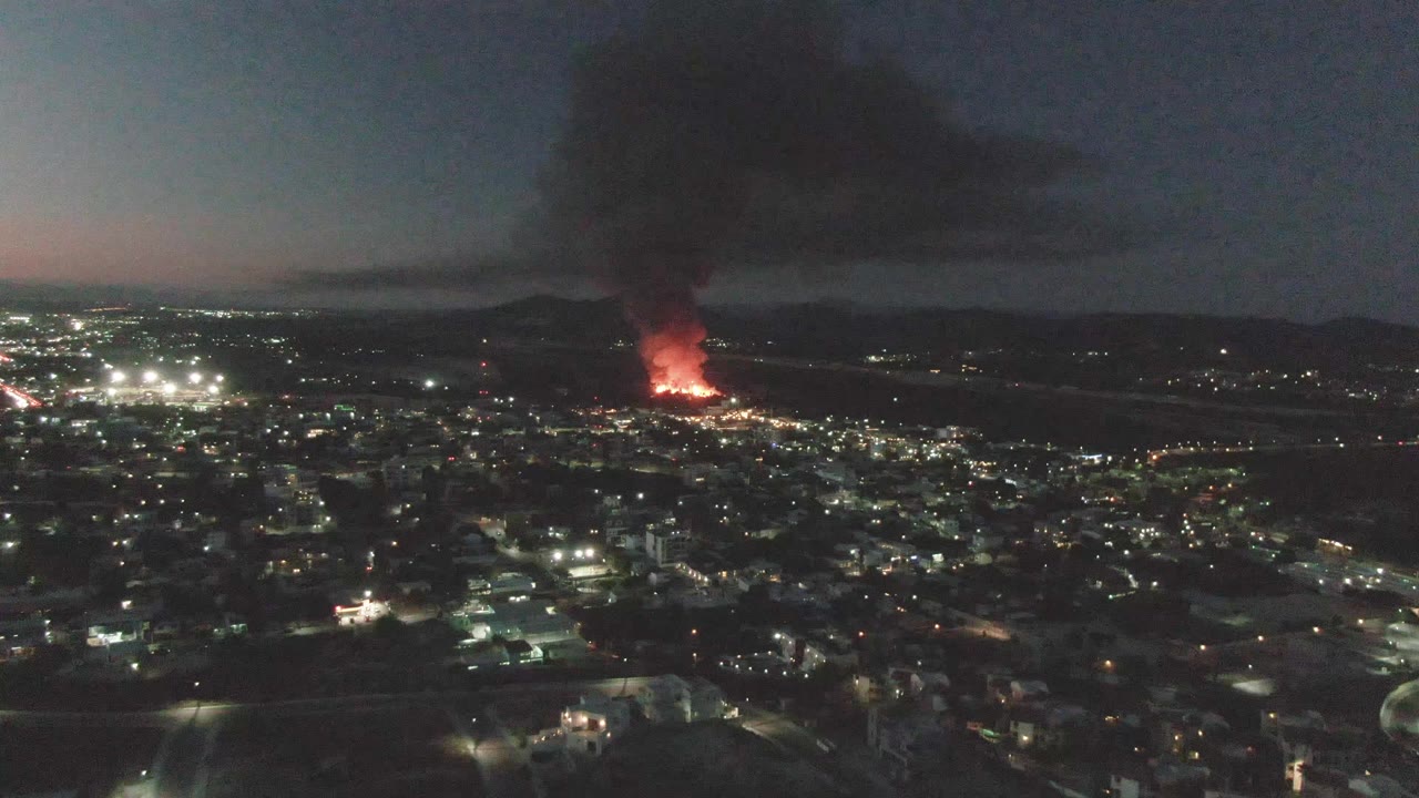 At 0:02, San Jose del Cabo appears as a broad nighttime city view with the fire glowing red near the horizon and a dense smoke column above it, with no visible people.