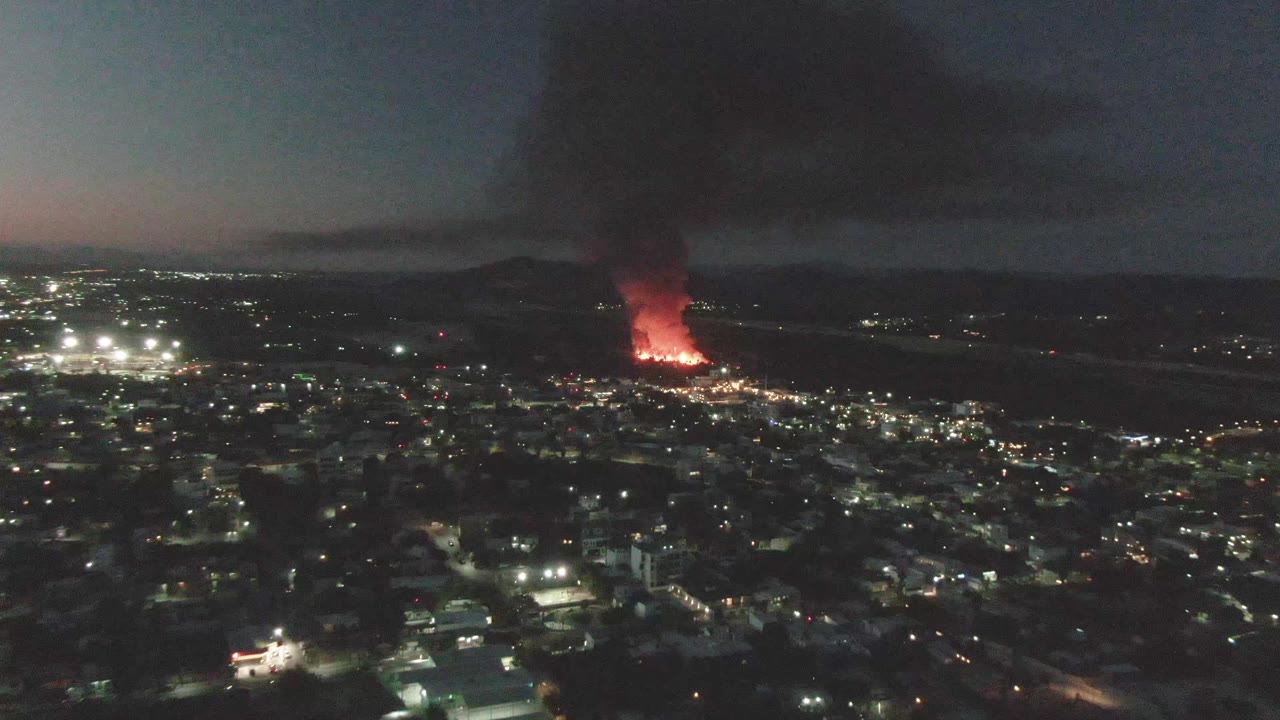 Wide aerial night view over San Jose del Cabo with a red fire glowing near the horizon, black smoke rising above city lights, and no visible people.