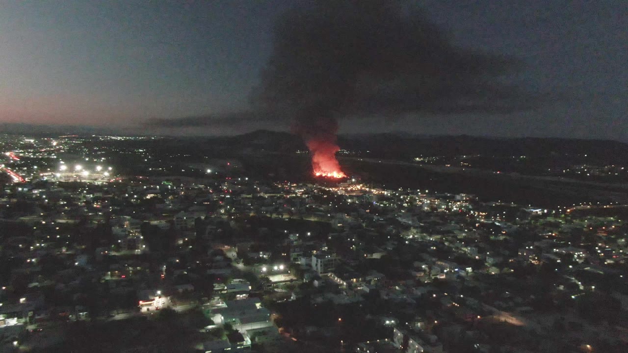 Aerial view of San Jose del Cabo at night with dense city lights below, a red fire burning in the distance, a thick smoke column above it, and no visible people.