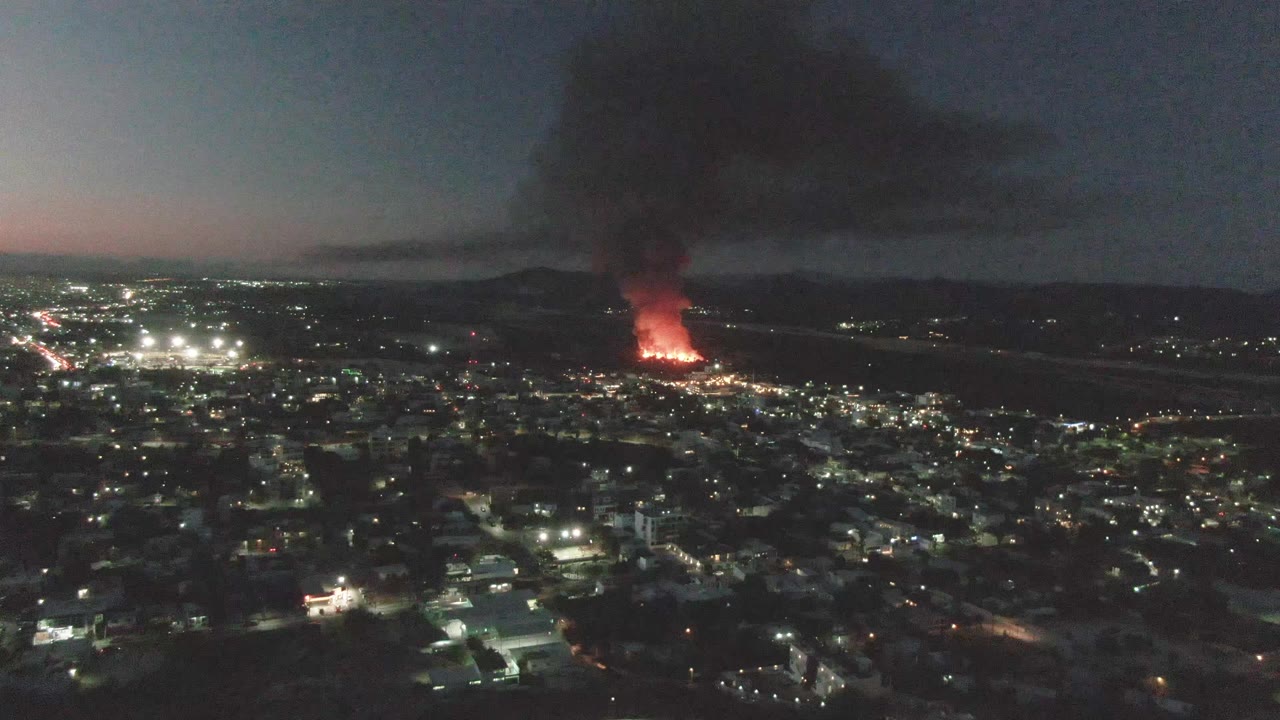 Night aerial panorama over San Jose del Cabo with illuminated streets and buildings leading toward a red fire, dark smoke spreading upward, and no visible people.