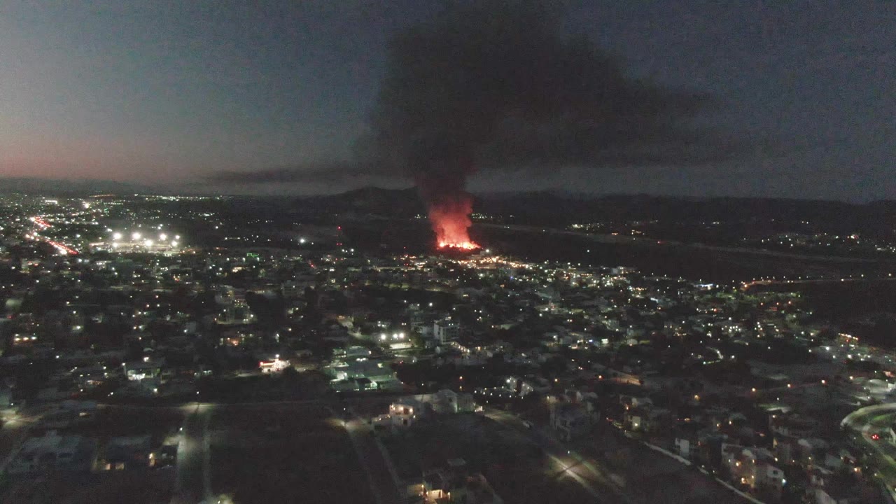 Aerial view over San Jose Del Cabo at dusk with city lights in the foreground, a red fire glowing in the distance, a black smoke plume rising, and no visible people.