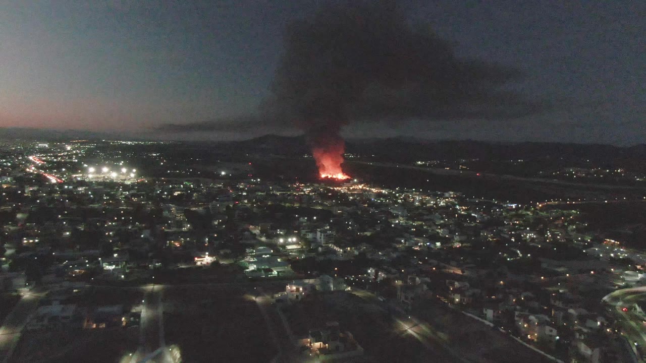 Elevated dusk city view with fire glow and smoke beyond roads and lights.